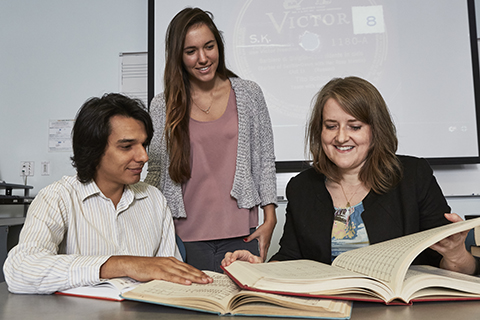 A teacher with long brown hair is turning the pages of a book while 2 students look on beside her