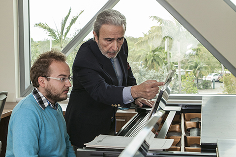 A man with white hair and beard wearing a dark suits points at a book propped up on a table while a man with glasses looks on intently.