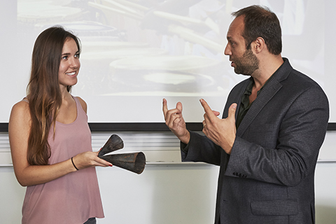 A woman in a pink top with long brown hair is holding up cowbells while a man in a dark suit motions with his hands