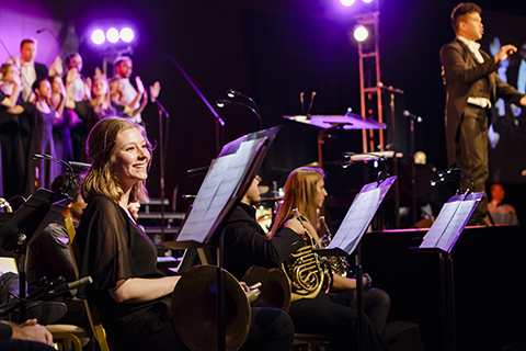 A woman with long blond hair and a black dress is sitting behind sheet music and smiling while a chorus sings in the background