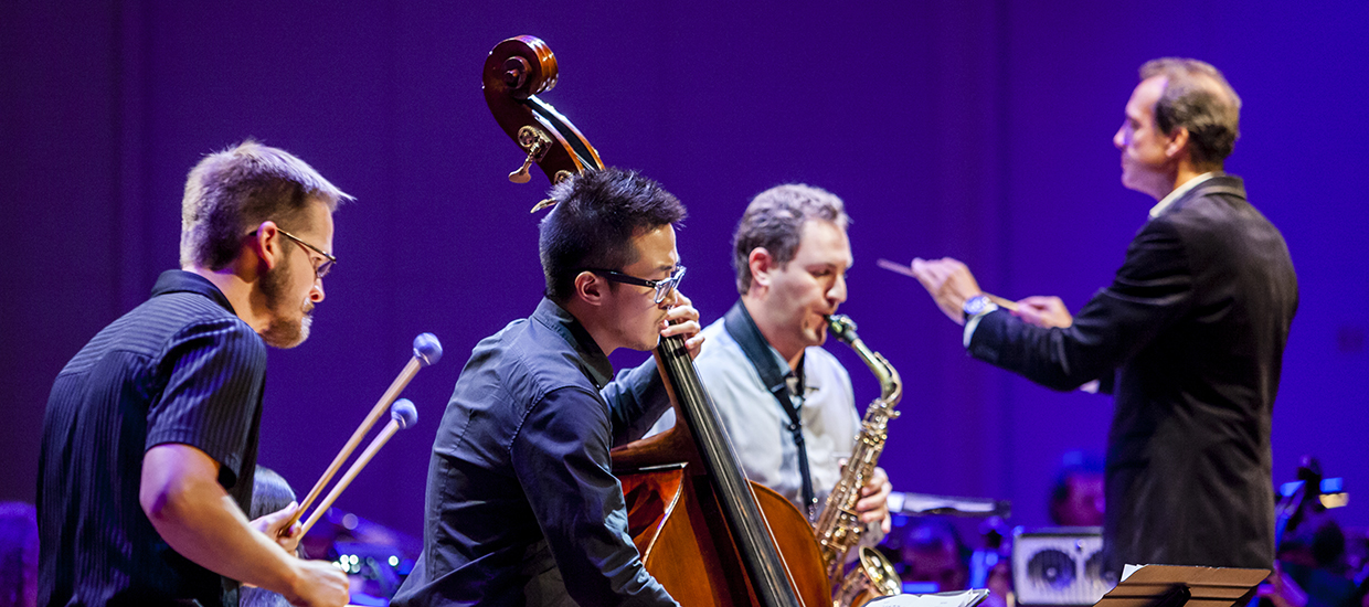 A group of musicians play the xylophone, upright bass, and saxophone as a conductor gestures with his baton in the background