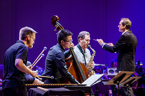 A group of musicians play the xylophone, upright bass, and saxophone as a conductor gestures with his baton in the background